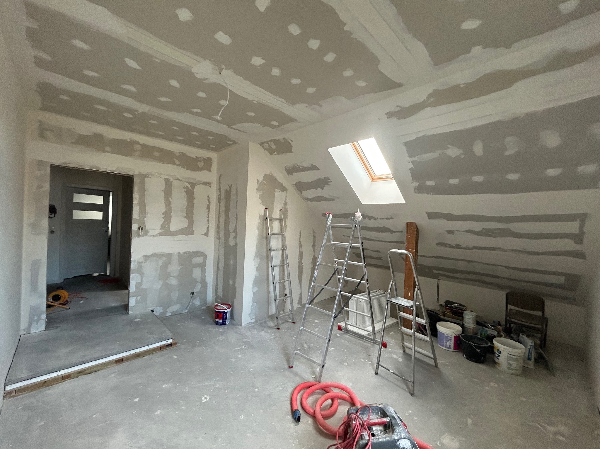Pokój na poddaszu w trakcie prac wykończeniowych: ściany i sufit z płyt karton-gips, widoczne łączenia szpachlowane, trzy drabiny aluminiowe, narzędzia i materiały budowlane na podłodze, okno dachowe.