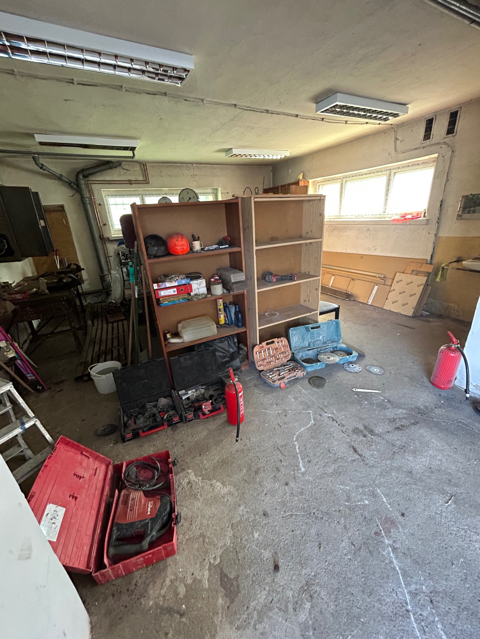 Warsztat z narzędziami, wiertarkami Hilti w czerwonych walizkach, regałami i gaśnicami, betonowa podłoga zarysowana kredą.