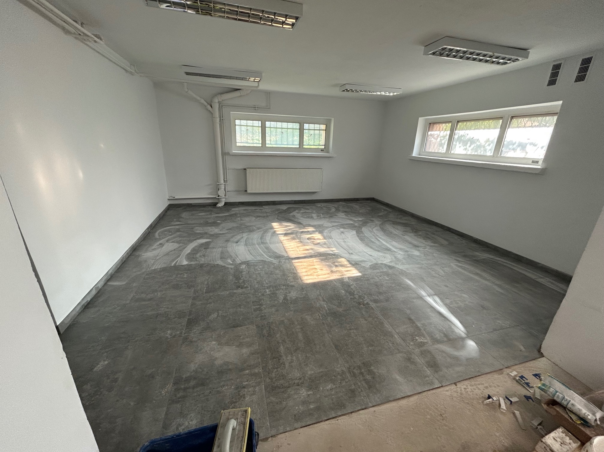 Szare płytki podłogowe po czyszczeniu w pustym, odnowionym pokoju z oknami i grzejnikiem, widoczne narzędzia glazurnicze.