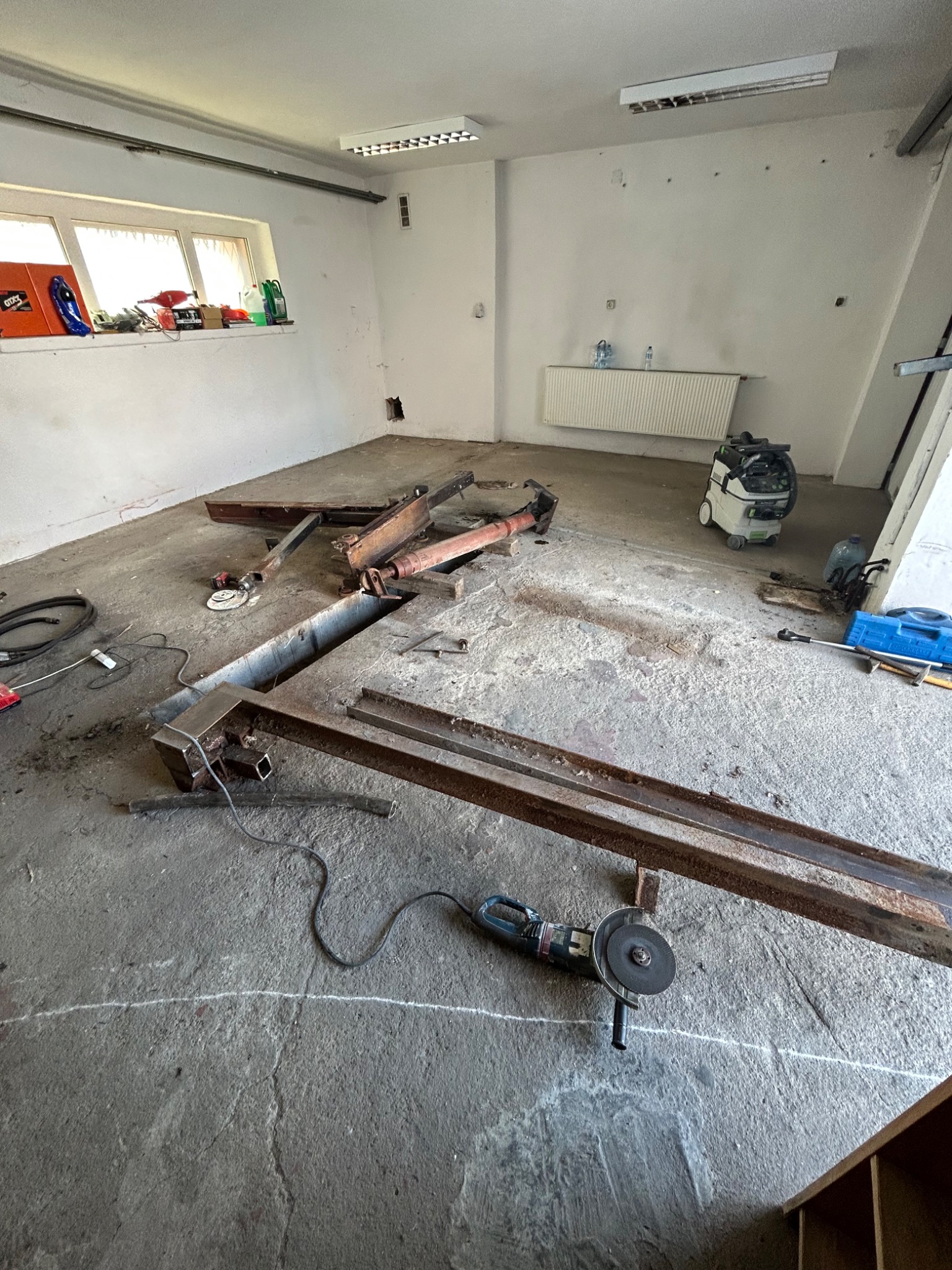 Wnętrze warsztatu w trakcie remontu, widoczny kanał naprawczy, elementy konstrukcyjne, szlifierka kątowa, odkurzacz przemysłowy i narzędzia.