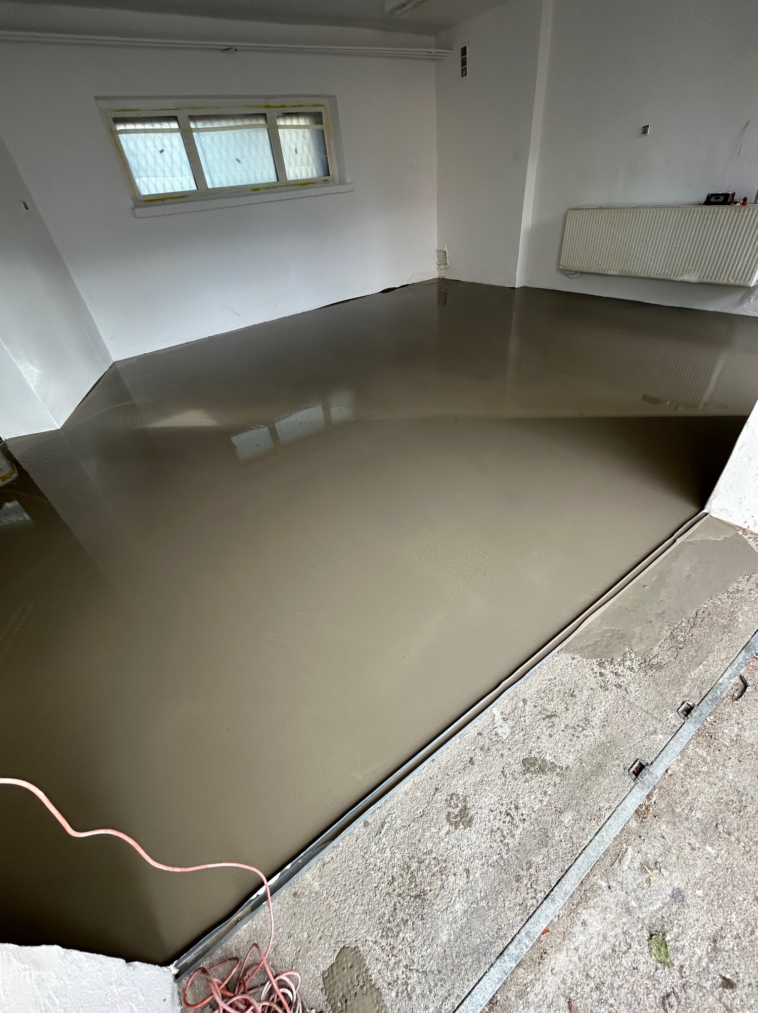 Wylewka samopoziomująca na podłodze w jasnym pomieszczeniu, widoczne kable i listwa dylatacyjna przy ścianie.