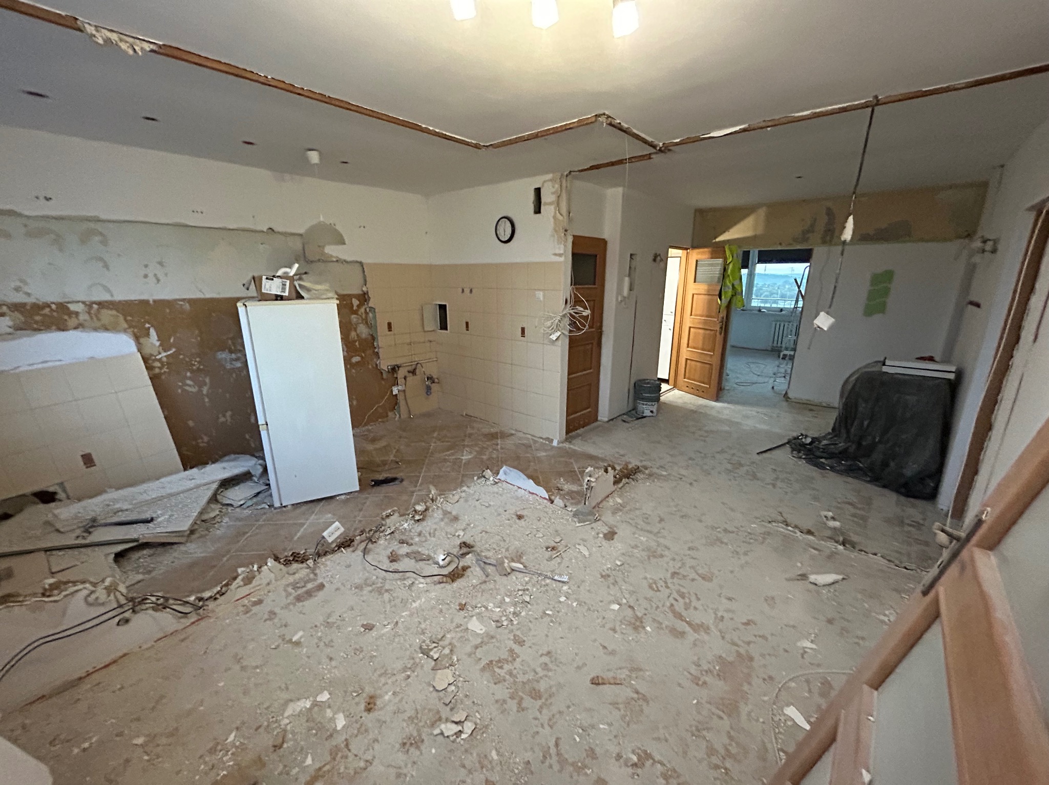 Wnętrze mieszkania w trakcie demontażu: zerwane płytki podłogowe, częściowo usunięte płytki ścienne w kuchni, odsłonięte przewody elektryczne, widoczny stary parkiet i biała lodówka.