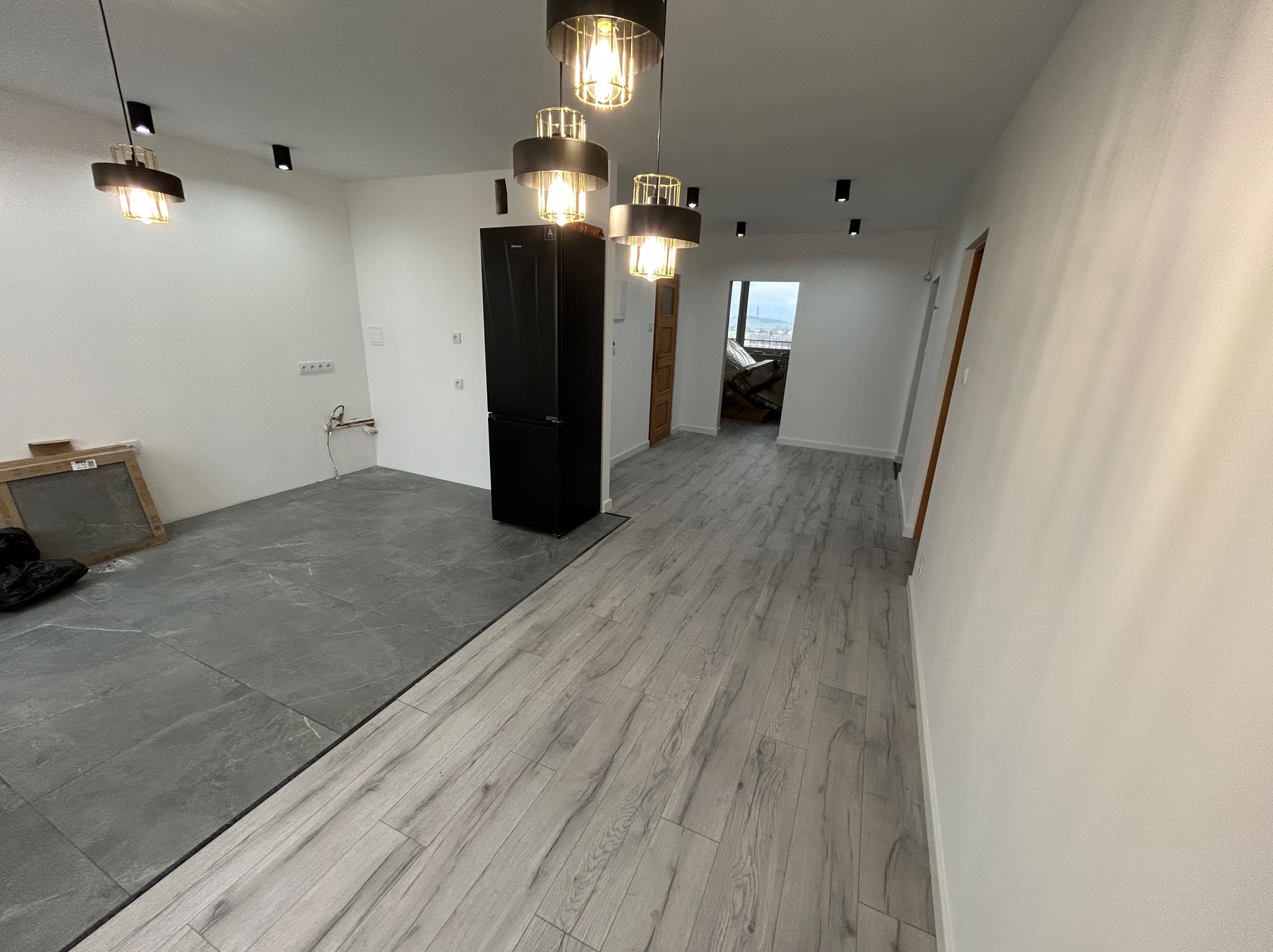 Wnętrze mieszkania w trakcie prac wykończeniowych: szare płytki podłogowe przechodzą w panele podłogowe imitujące drewno, białe ściany, czarna lodówka i nowoczesne lampy wiszące.