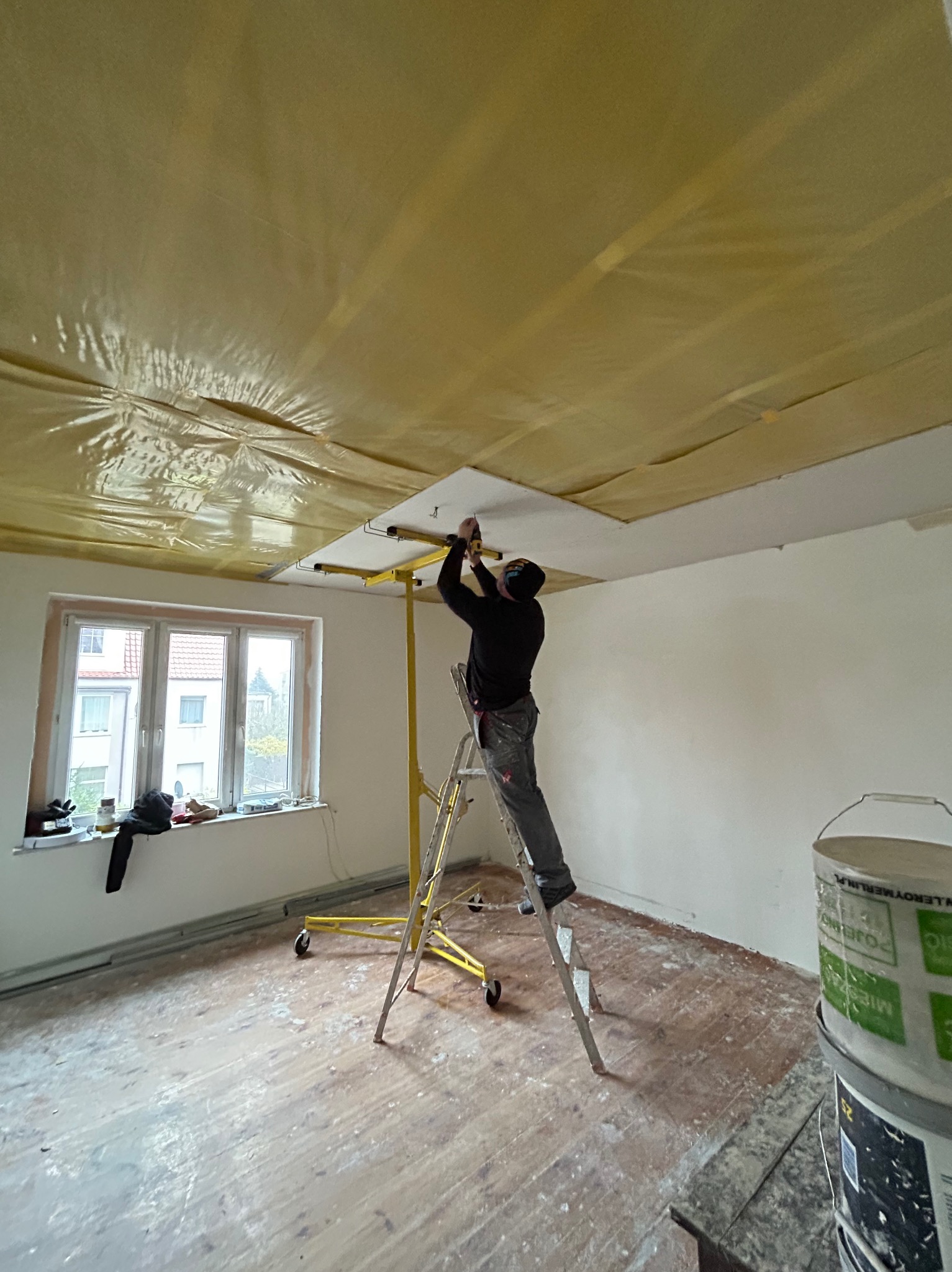 Pracownik na drabinie montuje płytę kartonowo-gipsową do sufitu z folią ochronną, używając podnośnika, widoczny bałagan w pokoju i wiadro farby.