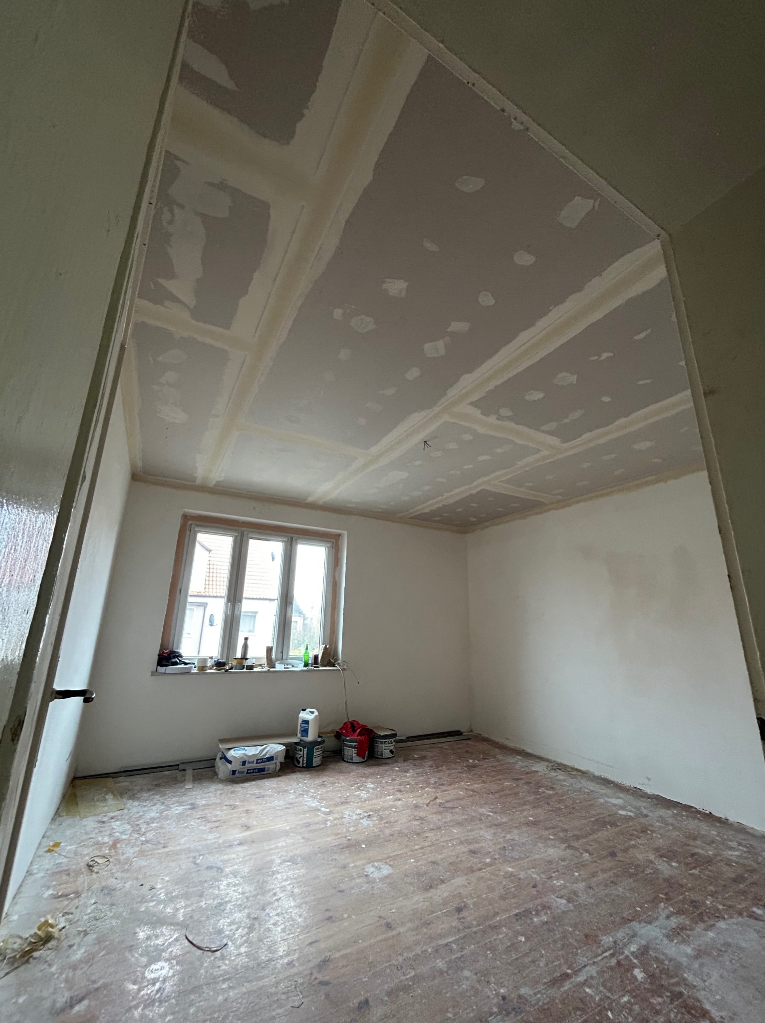 Pokój w trakcie remontu: sufit z płyt kartonowo-gipsowych z widocznymi łączeniami, przygotowany do dalszych prac wykończeniowych, stare drewniane podłogi, okno z widokiem na domy, materiały...