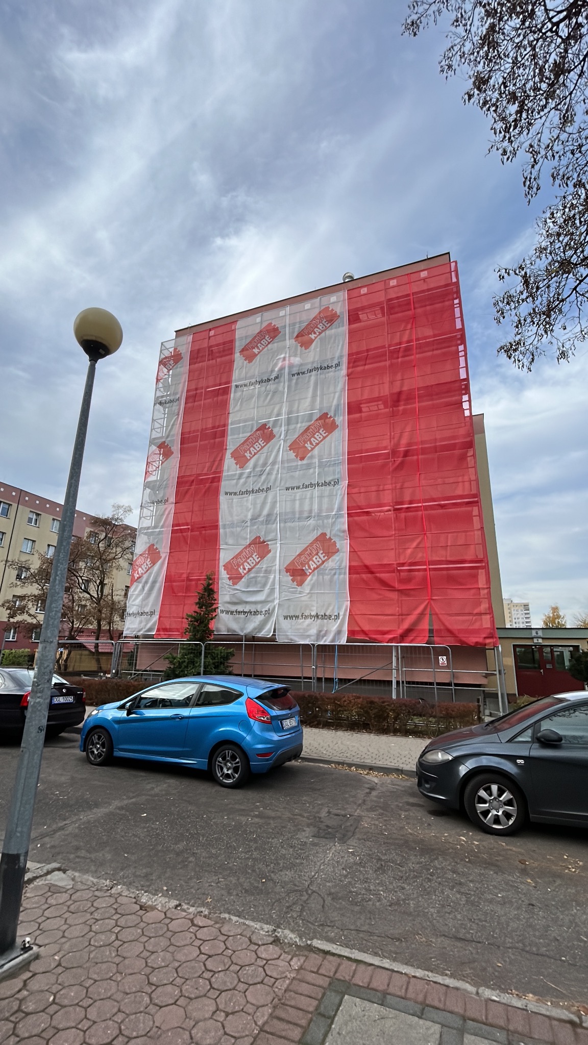 Budynek wielorodzinny w trakcie prac remontowych elewacji, osłonięty siatką z reklamami farb, widok z chodnika z zaparkowanymi samochodami.
