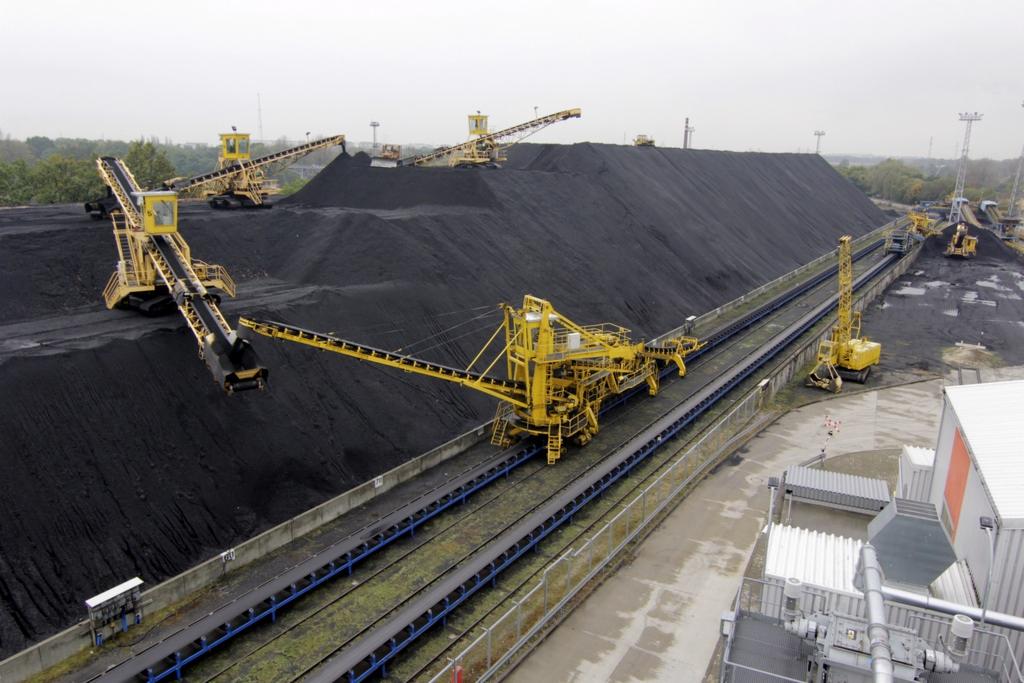 Duży pryzmatyczny stos czarnego miału węglowego z żółtymi maszynami transportującymi na szynach i taśmociągach, widok z góry.