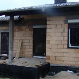 PHU Budoplast - Dom w trakcie budowy: wstawione brązowe drzwi zewnętrzne z dekoracyjnym wzorem i okno z roletą zewnętrzną, widoczne pustaki i fundamenty.