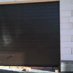 Ciemnobrązowa brama garażowa segmentowa, z poziomymi przetłoczeniami, zamontowana w surowym murze z białych bloczków betonowych.