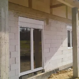 Nowe okno z roletą zewnętrzną w trakcie montażu w surowym budynku z betonu komórkowego, widoczne żółte rury instalacyjne na zewnątrz.