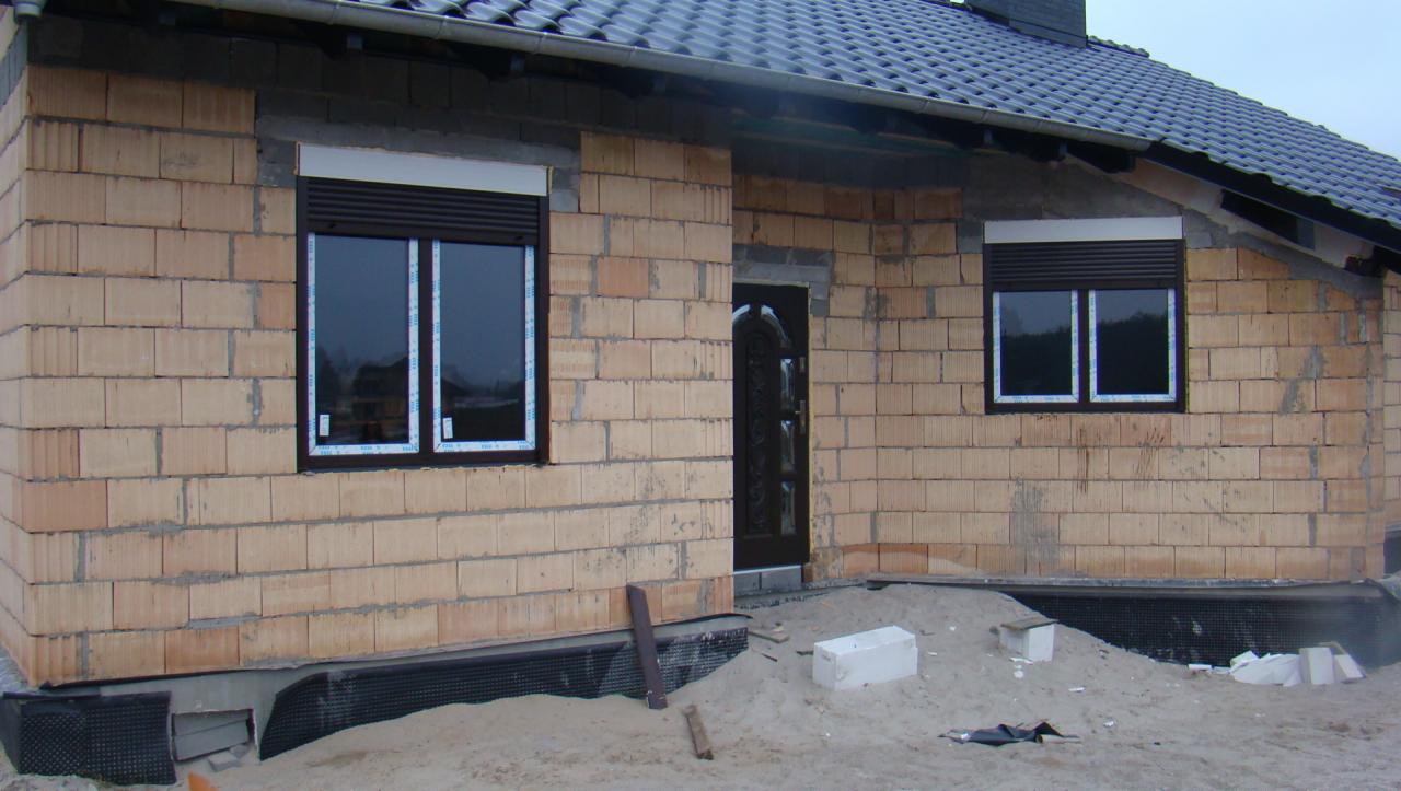 Surowy stan elewacji domu z zamontowanymi oknami PCV z brązowymi roletami zewnętrznymi i drzwiami wejściowymi, widoczne bloczki z betonu komórkowego i piasek.