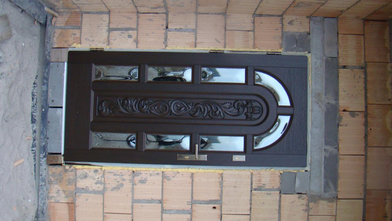 Brązowe drzwi wejściowe z dekoracyjnym wzorem i szybami, w trakcie montażu, widoczne cegły i pianka montażowa.