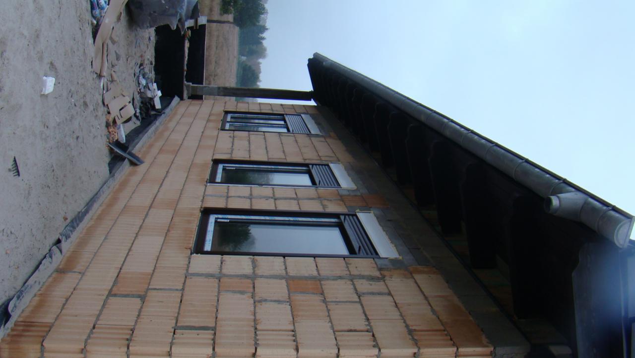 Budynek w trakcie budowy z zamontowanymi oknami z brązowymi ramami i zewnętrznymi roletami, widoczne pustaki ceramiczne i fragment dachu z rynną.