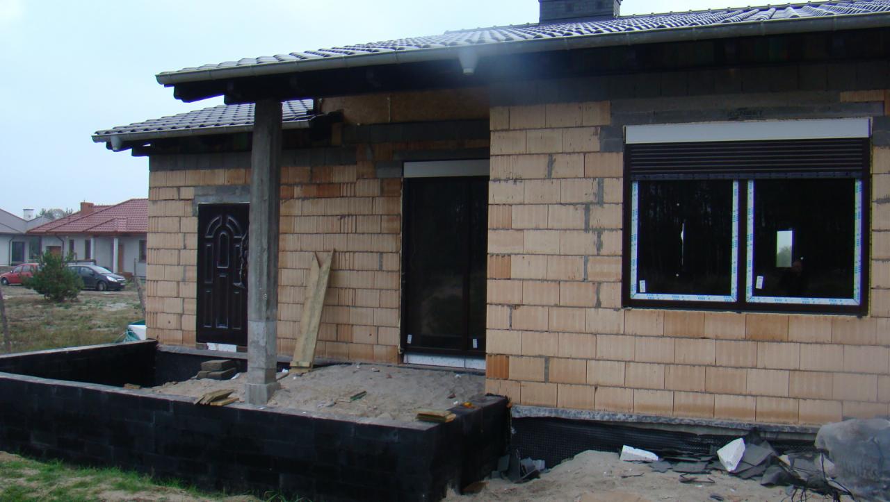 Dom w trakcie budowy: wstawione brązowe drzwi zewnętrzne z dekoracyjnym wzorem i okno z roletą zewnętrzną, widoczne pustaki i fundamenty.