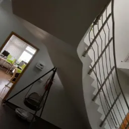 Klatka schodowa z krętymi schodami z metalową balustradą i widokiem na biuro przez otwarte drzwi, z wiszącym fotelem obok schodów.