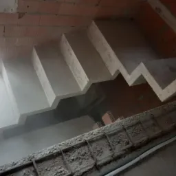 Surowe, betonowe schody w trakcie budowy, widok z góry na zbrojenie i ceglaną ścianę w tle.