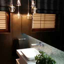 Nowoczesna łazienka z czarnymi płytkami, lustrem z mozaiką, białym blatem z umywalką i dekoracyjną rośliną w koszyku, oświetlona trzema wiszącymi lampami z metalowymi kloszami.