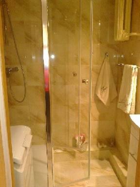 Wnętrze łazienki z kabiną prysznicową z przezroczystymi drzwiami, wyłożonej marmurkowymi płytkami w odcieniach beżu i brązu. Widoczny fragment pralki, ręczniki i kosmetyki.