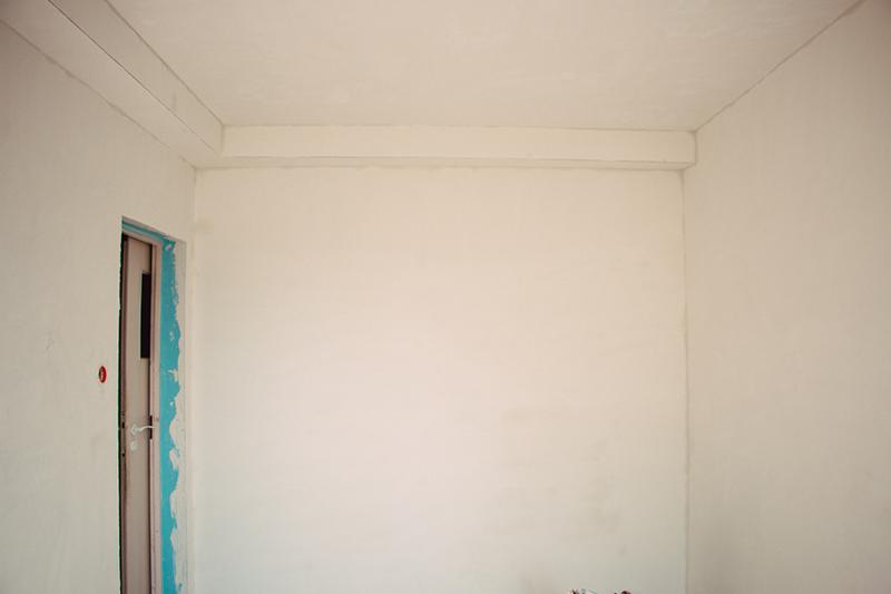 Wnętrze pokoju w trakcie prac wykończeniowych, widoczne ściany po nałożeniu warstwy gładzi, listwa pod sufitem i futryna drzwi zabezpieczona taśmą malarską.