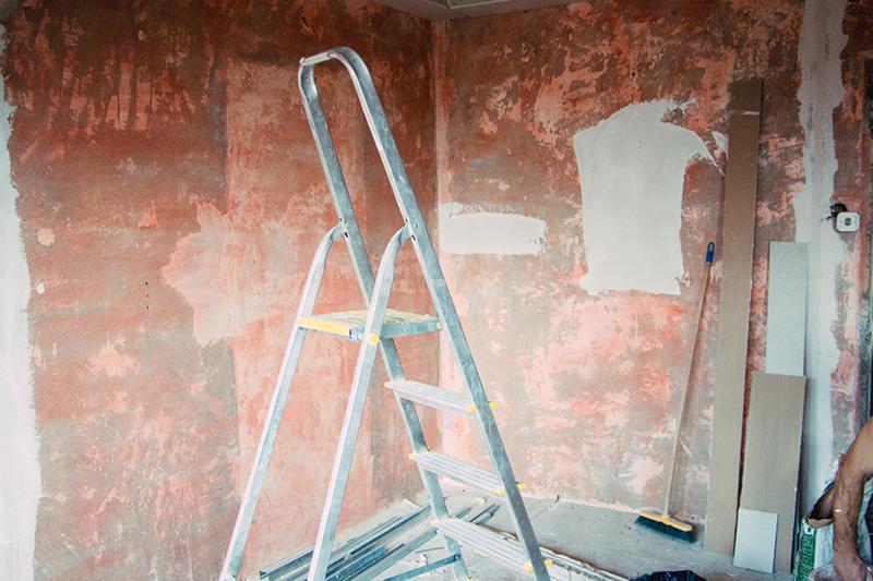 Pokój w trakcie przygotowania do malowania ścian, widoczna drabina aluminiowa, ściany z pozostałościami starej farby i fragmentami nałożonej gładzi, listwy przypodłogowe oparte o ścianę.