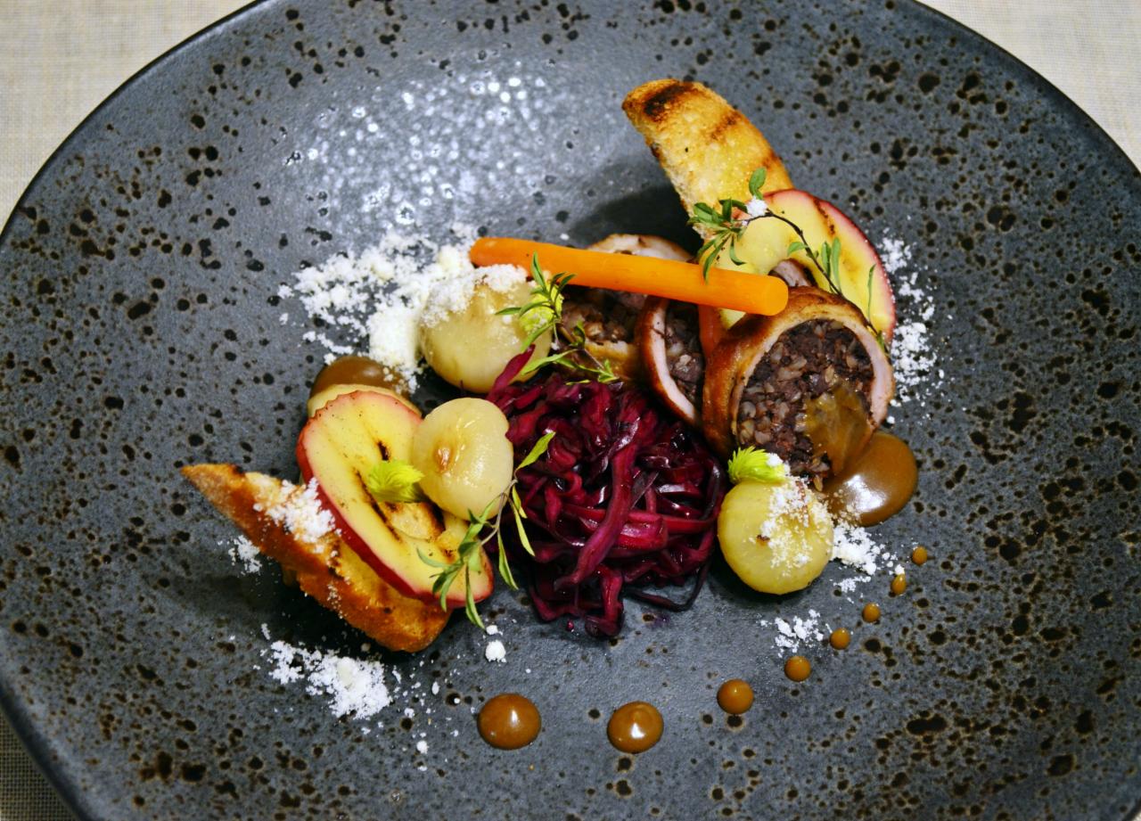 Elegancko podane danie na ciemnym, nakrapianym talerzu: kaszanka zawinięta w mięso, czerwona kapusta, karmelizowana cebula, grillowana jabłko, grzanka, marchewka, posypane białym proszkiem i polane...