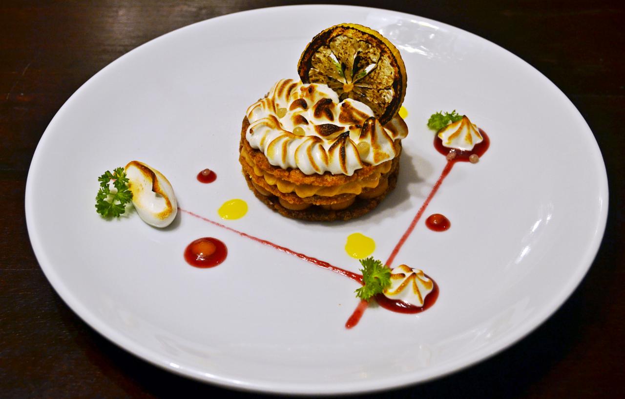 Elegancko udekorowany deser na białym talerzu: warstwowe ciasto z kremem, zwieńczone opiekaną cytryną i kleksami z sosem owocowym i bezą.