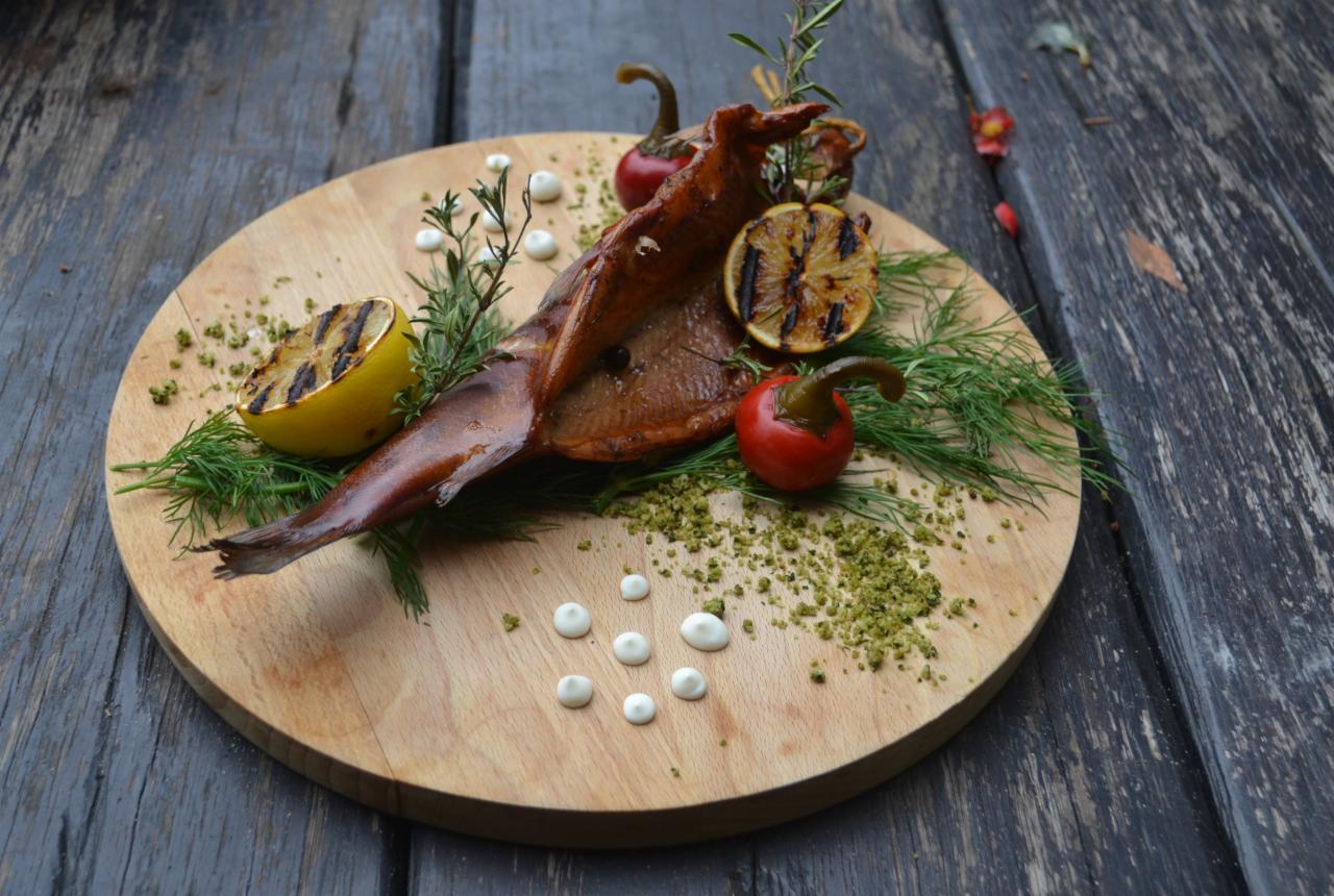 Wędzona ryba na drewnianej desce, udekorowana grillowaną cytryną, czerwoną papryką, koperkiem, rozmarynem, zielonymi przyprawami i kropkami sosu.