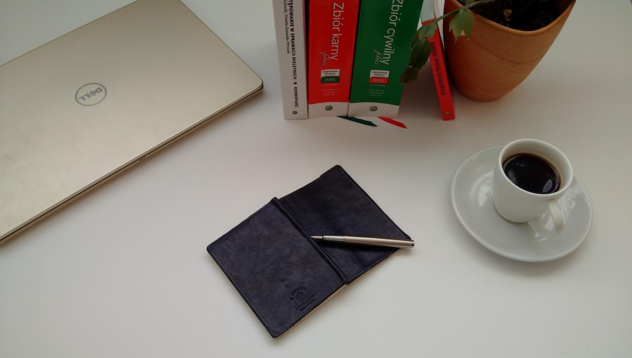 Biurko z laptopem Dell, notatnikiem ze srebrnym długopisem, kubkiem czarnej kawy i książkami prawniczymi Zbiór Karny i Zbiór Cywilny oraz roślina w doniczce.