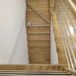 Widok z góry na jasne, drewniane schody z metalową balustradą, częściowo zamontowane, z widocznym narzędziem przy ścianie.