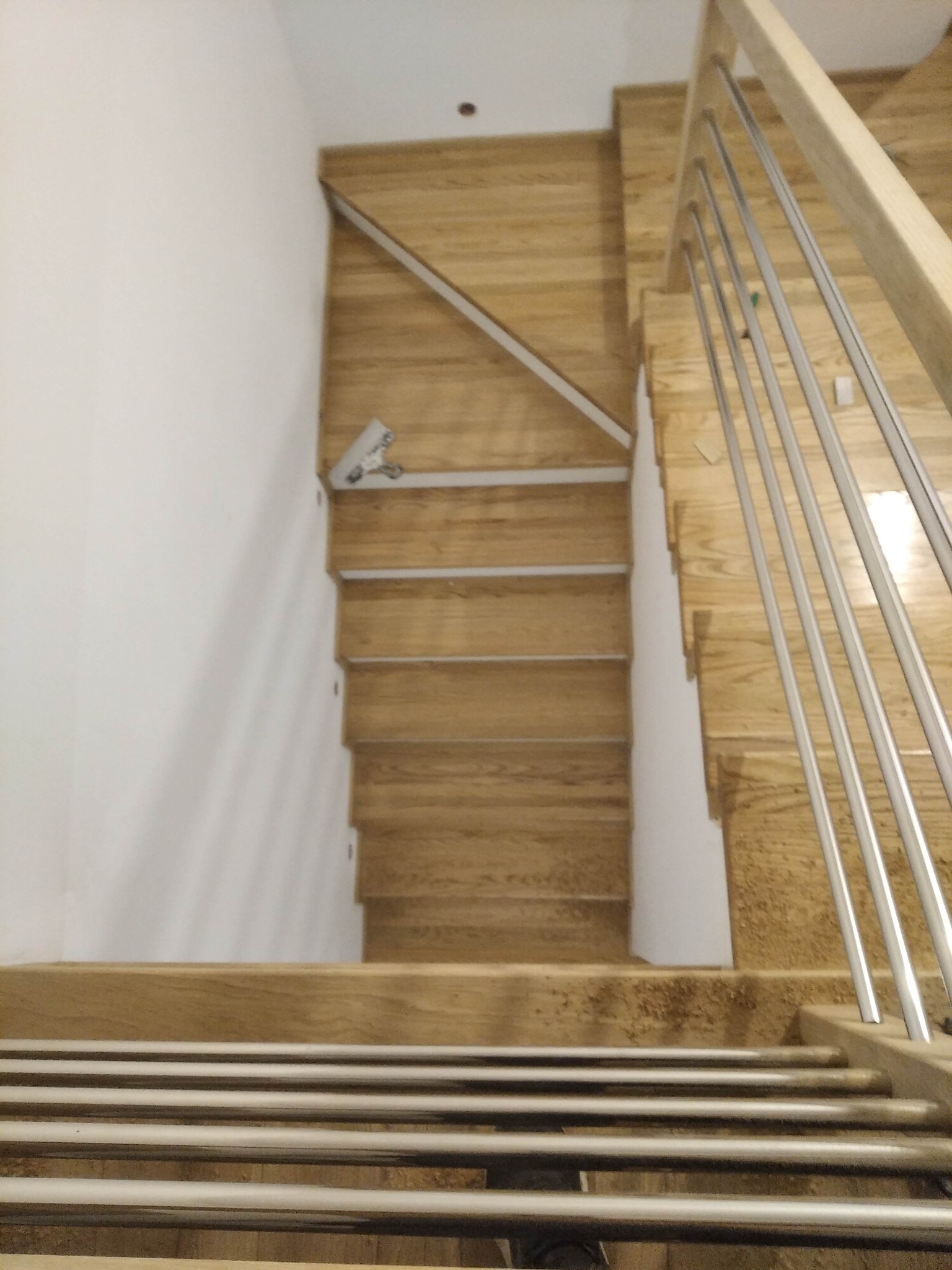 Widok z góry na jasne, drewniane schody z metalową balustradą, częściowo zamontowane, z widocznym narzędziem przy ścianie.