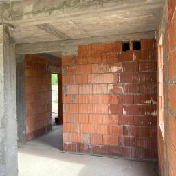 KAMIL KUCHARCZYK CONCRETE - Wnętrze surowego domu w trakcie budowy z widocznymi ścianami z czerwonej cegły ceramicznej oraz betonowym stropem i słupami nośnymi. Widoczne otwory okienne i drzwiowe.