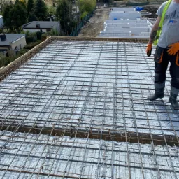 Przygotowanie zbrojenia stropu na budowie domu jednorodzinnego, widoczne siatki zbrojeniowe i pracownicy w odzieży roboczej.