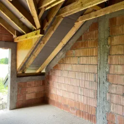 Wnętrze surowego domu w trakcie budowy, widoczne ściany z czerwonej cegły, drewniana konstrukcja dachu i wylewka podłogowa. Przez otwór okienny widać zieleń na zewnątrz.