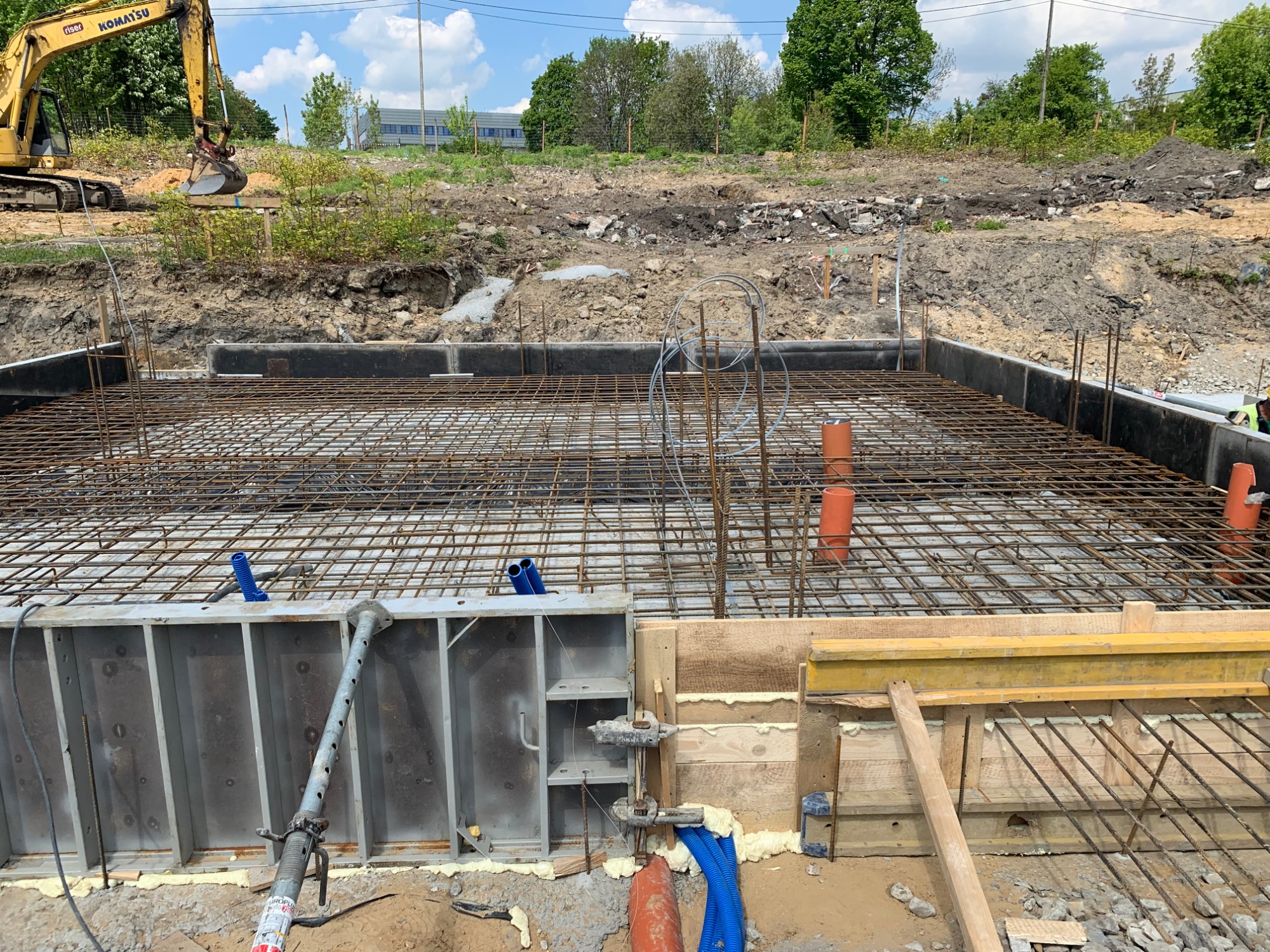 Siatka zbrojeniowa przygotowana do zalania betonem, widoczne szalunki metalowe i drewniane, pomarańczowe rury instalacyjne oraz koparka w tle.