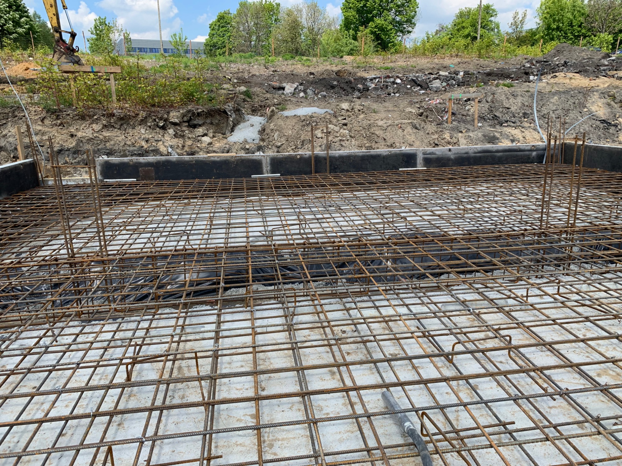 Zbrojenie fundamentu z widoczną siatką stalową i wystającymi prętami, przygotowane do wylania betonu, z ziemnym wykopem w tle.