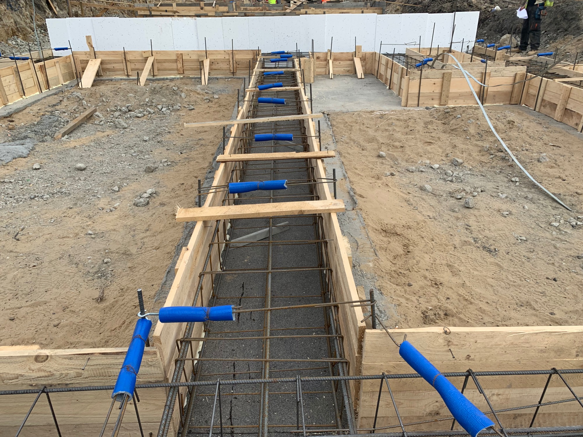 Szeroki kadr na wykop z drewnianym szalunkiem i siatką zbrojeniową, zabezpieczoną niebieską folią, przygotowany pod fundament, z widocznym terenem budowy w tle.