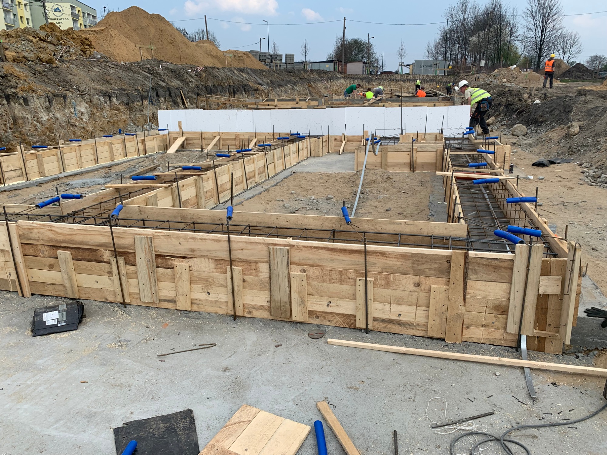 Przygotowanie szalunków i zbrojenia pod fundamenty domu jednorodzinnego, widoczni pracownicy w odblaskowych kamizelkach.