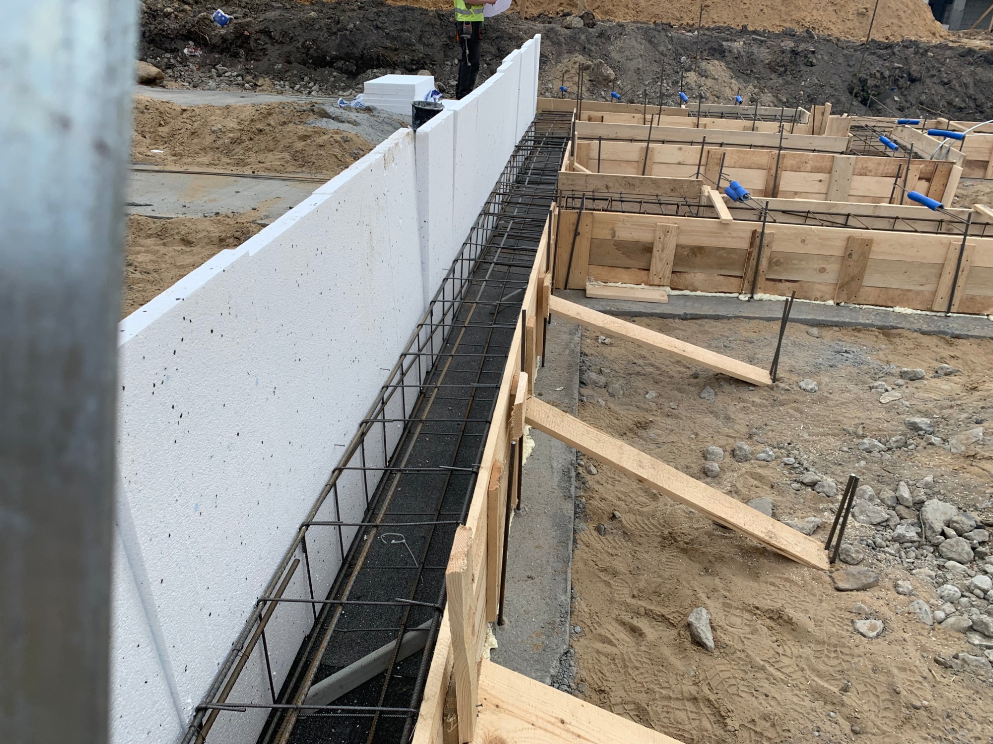 Przygotowanie szalunku i zbrojenia pod fundament, widoczne białe płyty izolacyjne pionowo, drewniane szalunki oraz stalowe pręty zbrojeniowe, w tle usypany grunt.