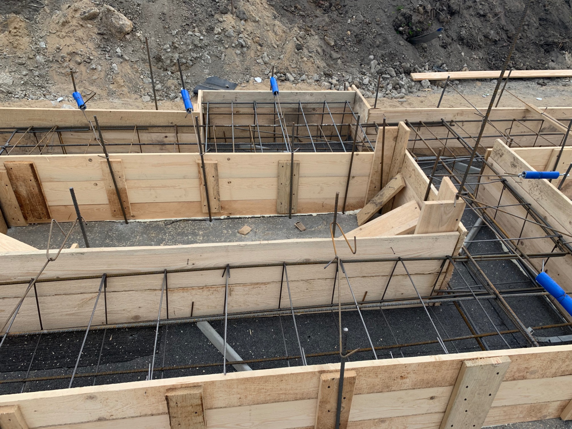 Szczegółowe ujęcie szalunku fundamentowego zbrojonego stalowymi prętami, zabezpieczonego niebieskimi elementami dystansowymi, przygotowanego do wylania betonu.