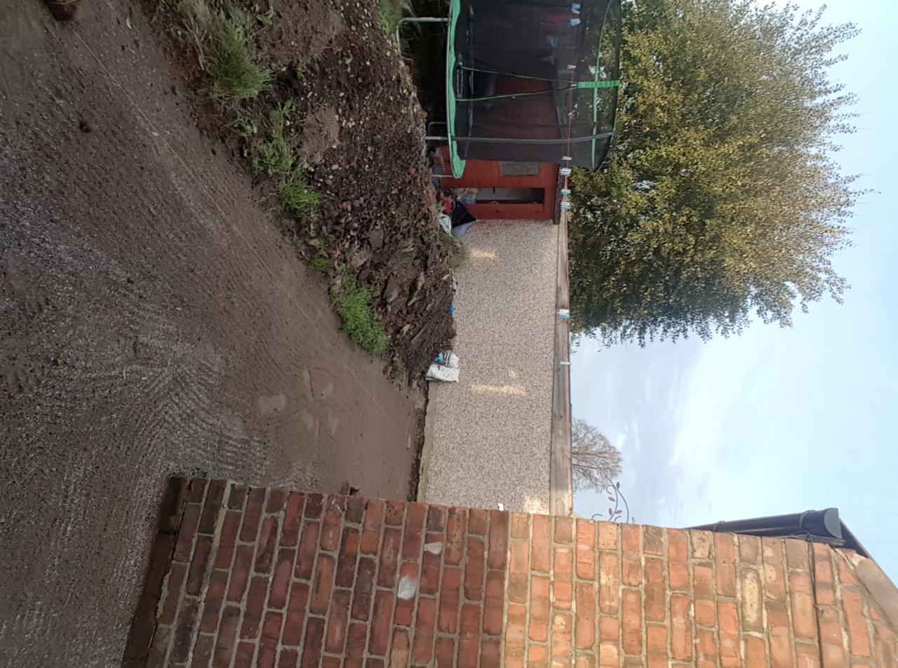 Przygotowanie terenu pod budowę: wykopana ziemia, widoczny fragment fundamentu, ceglany mur w tle, trampolina. Ziemia z wyraźnymi śladami opon.