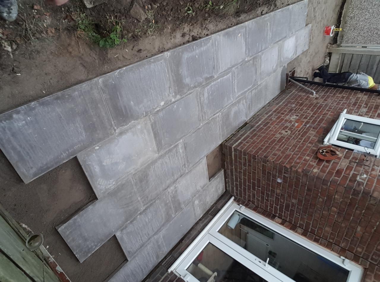 Szare, prostokątne płyty układane na ziemi, prawdopodobnie jako podstawa pod taras lub chodnik, częściowo przylegające do ceglanej ściany budynku z białymi oknami.