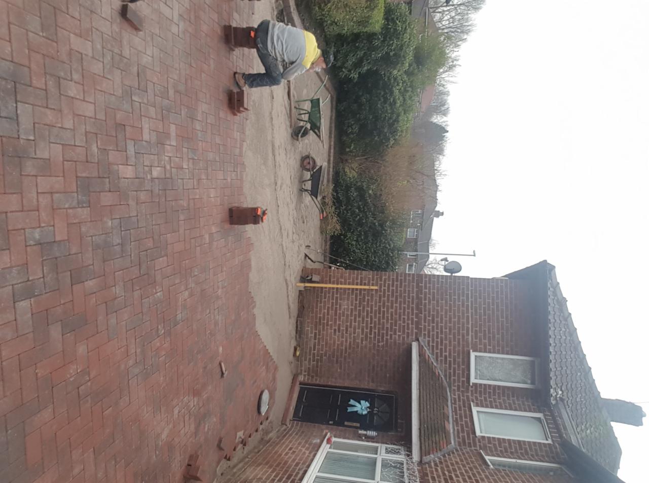 Pracownik w żółtej kamizelce układa czerwoną kostkę brukową na podjeździe przed domem z cegły, widoczne narzędzia i taczki.