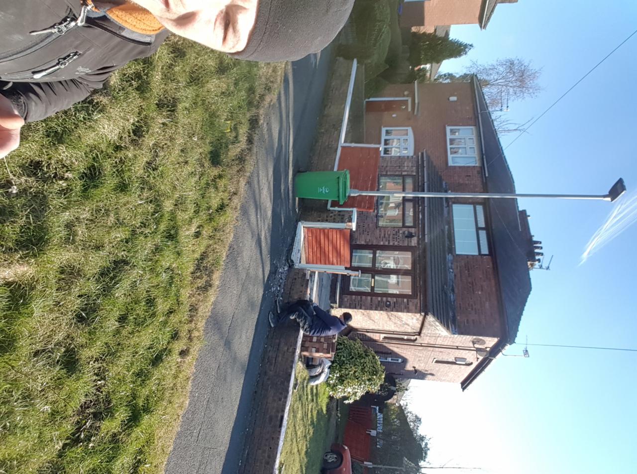 Pracownik w Manchesterze układa kostkę brukową na podjeździe przed domem z cegły, widoczny fragment trawnika i chodnika.