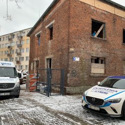 Izo Concept Sp. z o.o. - Budynek z czerwonej cegły w trakcie prac, z widocznym pracownikiem w oknie. Na pierwszym planie samochody firmy z logo. Zimowa sceneria, częściowo ośnieżona kostka brukowa.