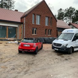 Izo Concept Sp. z o.o. - Nowoczesny dom w trakcie ocieplania pianką PUR, z czerwonym Mini Cooperem i firmowym vanem IzoConcept na błotnistym podwórku. Widoczne niedokończone ocieplenie.