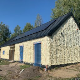 Izo Concept Sp. z o.o. - Budynek w trakcie izolacji: ściany pokryte pianą PUR, okna zabezpieczone folią. Widoczne panele słoneczne na dachu i fragment otoczenia z drzewami. Słoneczny dzień.