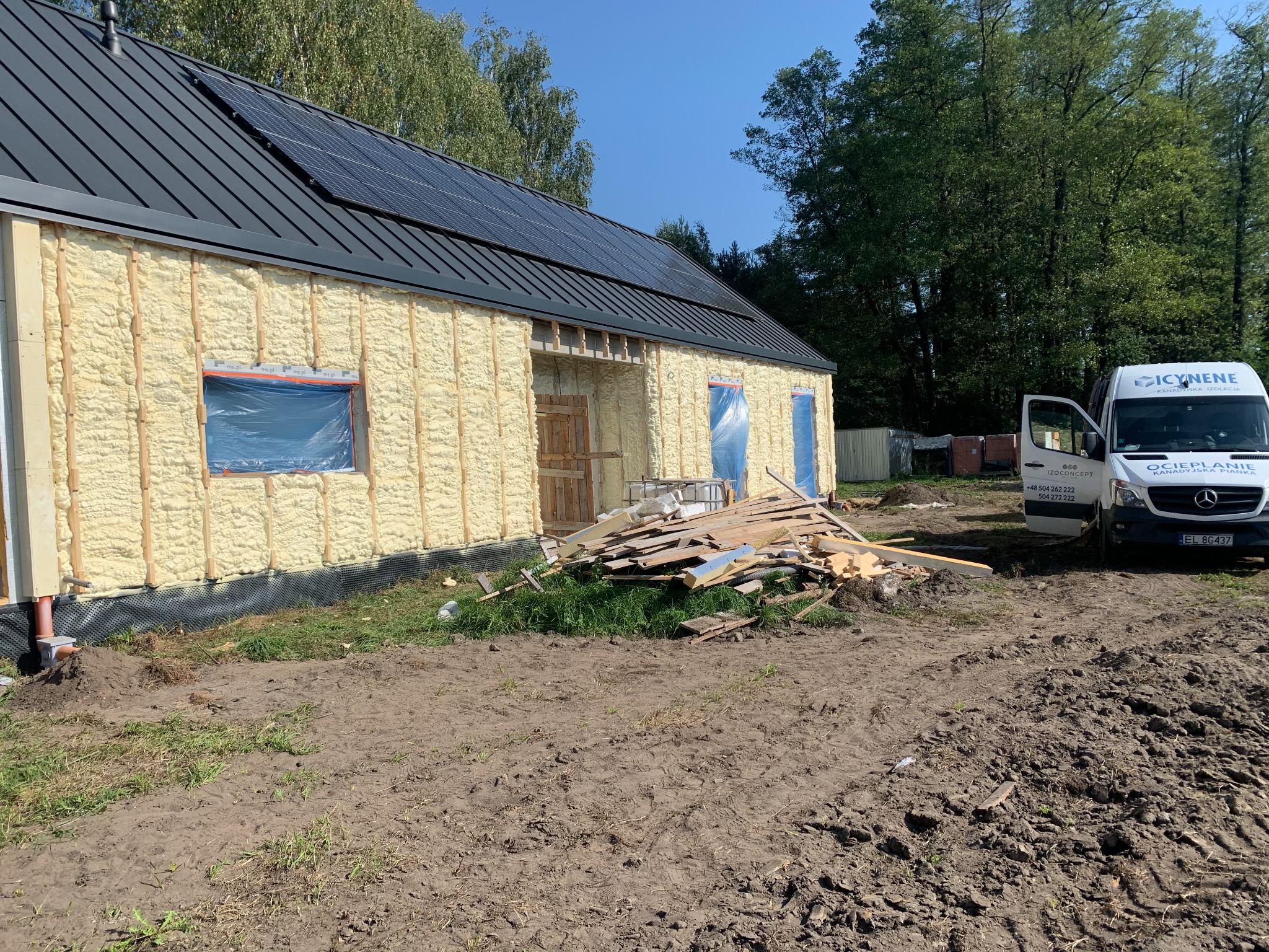 Ocieplony pianką PUR budynek z panelami słonecznymi na dachu. Widoczny firmowy bus i sterta drewna. Okna zabezpieczone folią. Prace w toku, teren wokół rozkopany.