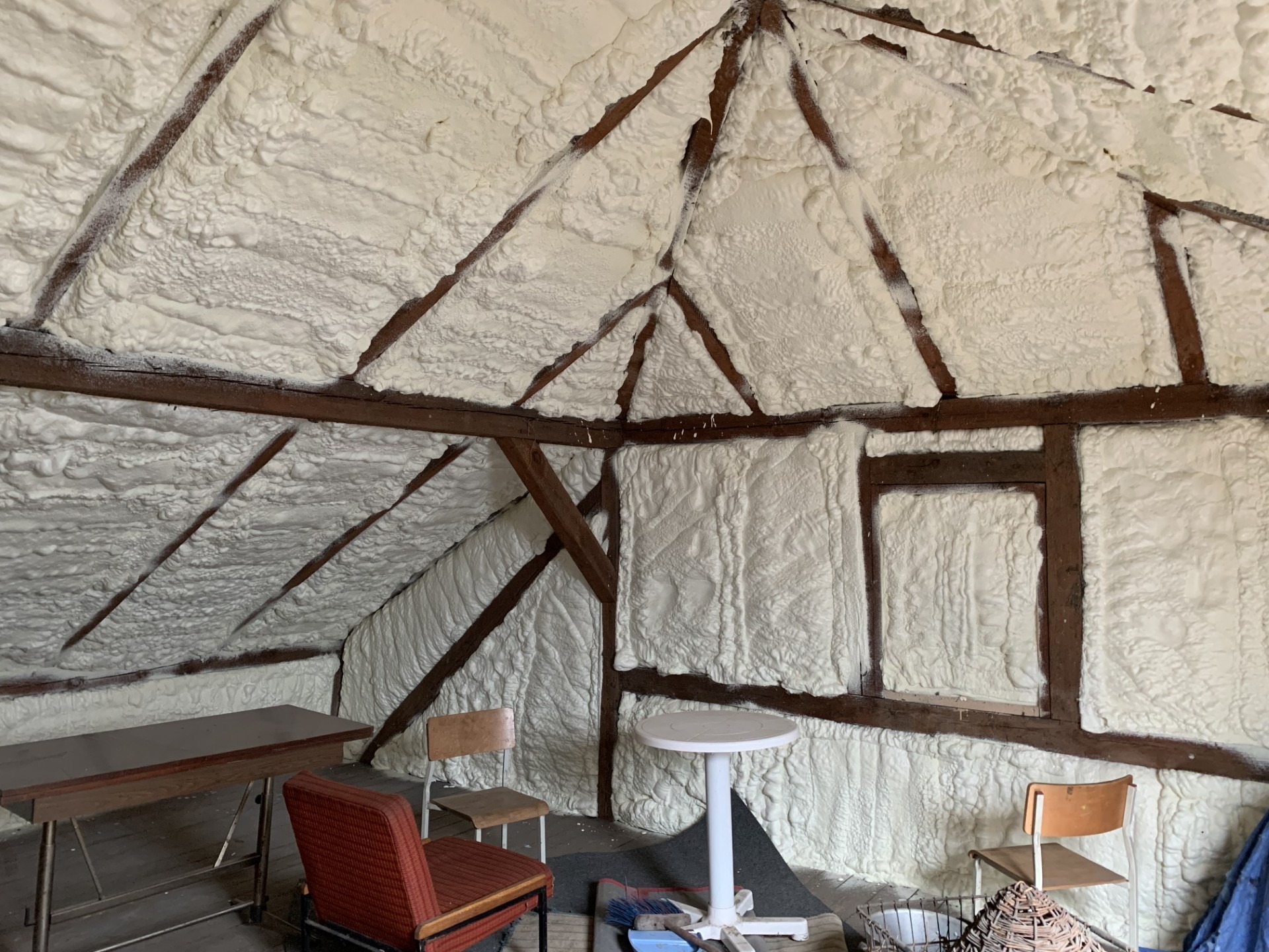 Poddasze po natrysku izolacji: biała pianka szczelnie wypełnia przestrzeń między drewnianymi belkami, tworząc jednolitą warstwę izolacyjną. Widoczne meble w pomieszczeniu.