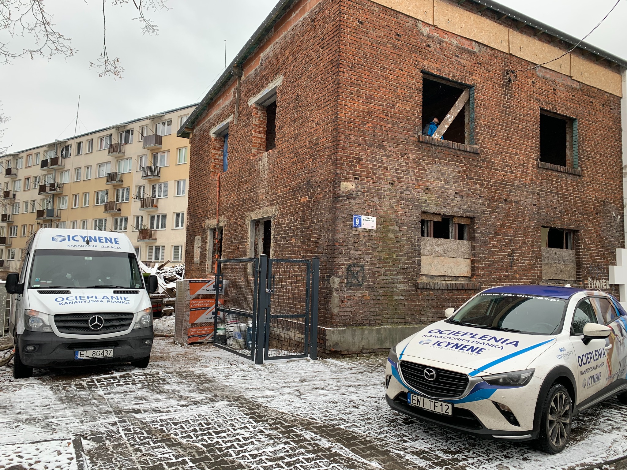 Budynek z czerwonej cegły w trakcie prac, z widocznym pracownikiem w oknie. Na pierwszym planie samochody firmy z logo. Zimowa sceneria, częściowo ośnieżona kostka brukowa.