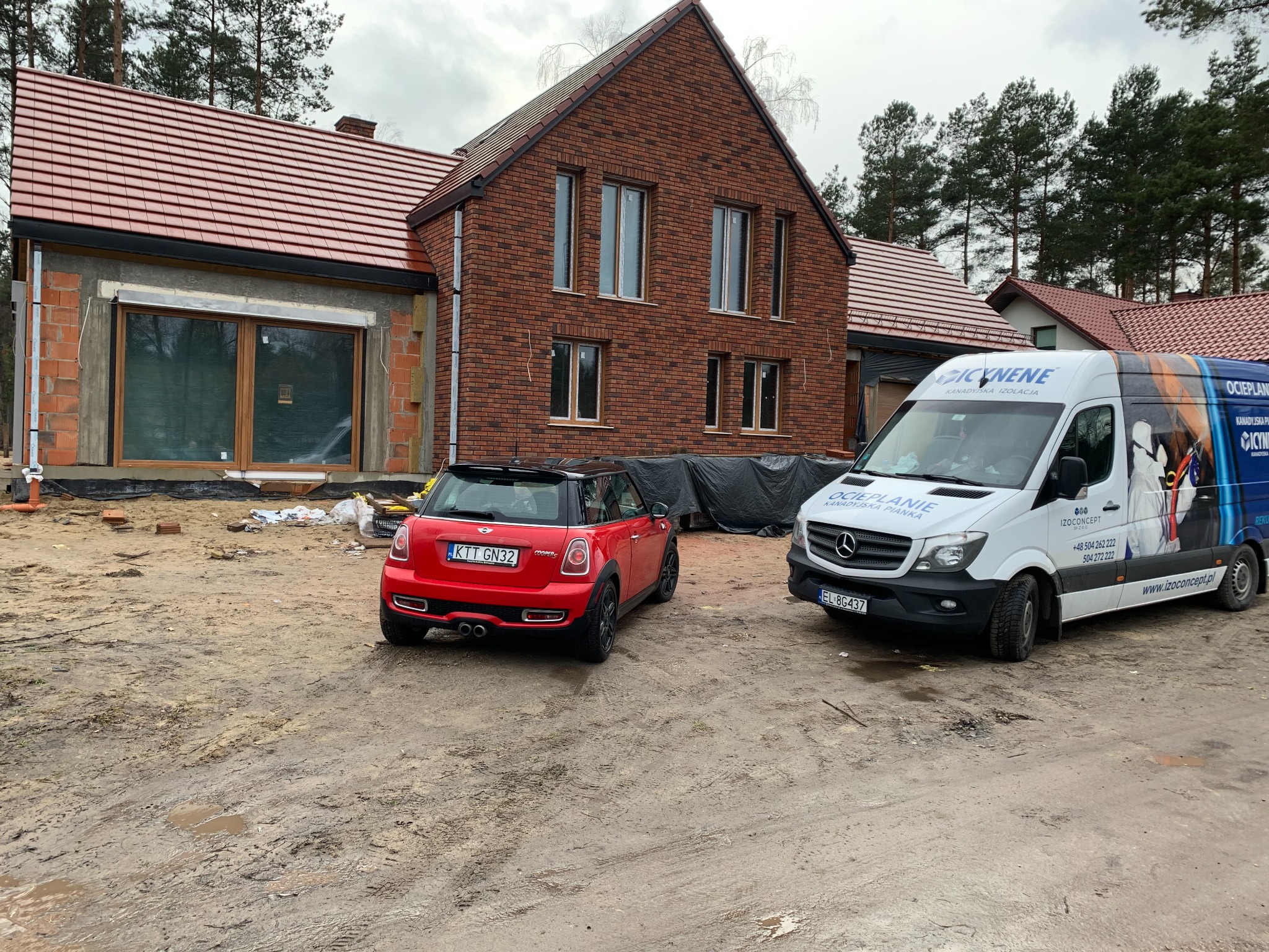 Nowoczesny dom w trakcie ocieplania pianką PUR, z czerwonym Mini Cooperem i firmowym vanem IzoConcept na błotnistym podwórku. Widoczne niedokończone ocieplenie.